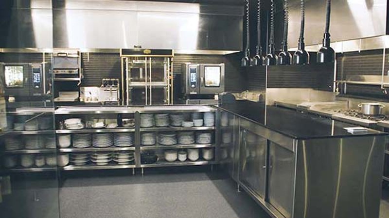 Professional stainless steel kitchen with ovens, shelves of plates, and prep counters.