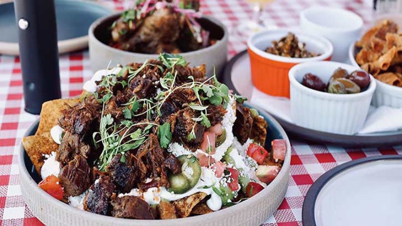 A variety of Mediterranean dishes on a red and white checkered tablecloth, including a large bowl of nachos topped with meat, vegetables, and sauce, accompanied by side bowls of olives and dips.