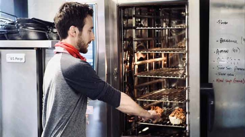 Person checking trays of baked goods in an industrial kitchen oven, with a list of ingredients written on a whiteboard in the background.