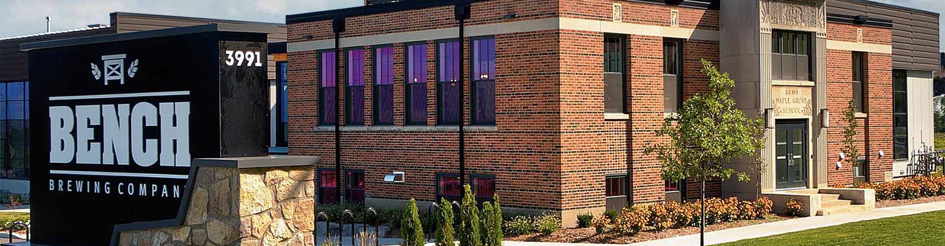 Exterior view of Bench Brewing Company with a prominent logo on a sign, located in a modern brick building with large windows and manicured landscaping.