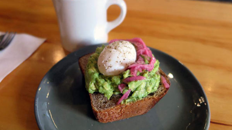 A plate of avocado toast topped with a poached egg and pickled onions, served with a cup of coffee.