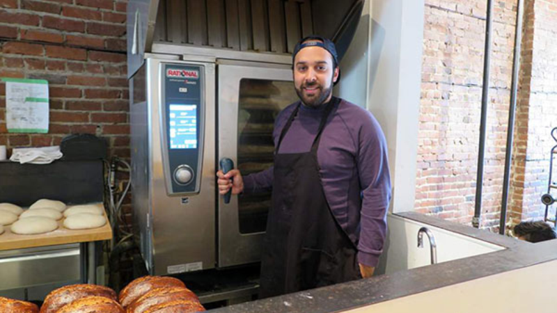 A baker stands in front of an oven, holding a tray, with freshly baked bread visible.