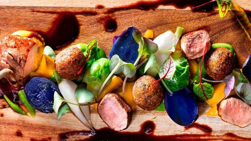 A beautifully plated dish with meat, vegetables, and sauce drizzled on a wooden board.