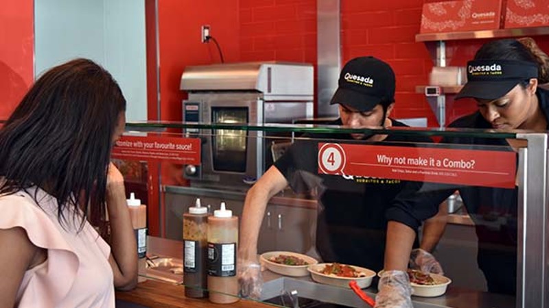 A customer at the counter of a restaurant watching employees prepare food with condiments.