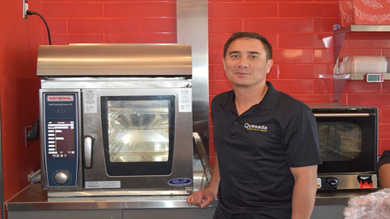 A man standing next to a Rational oven in a kitchen with a red wall, wearing a Quesada shirt.