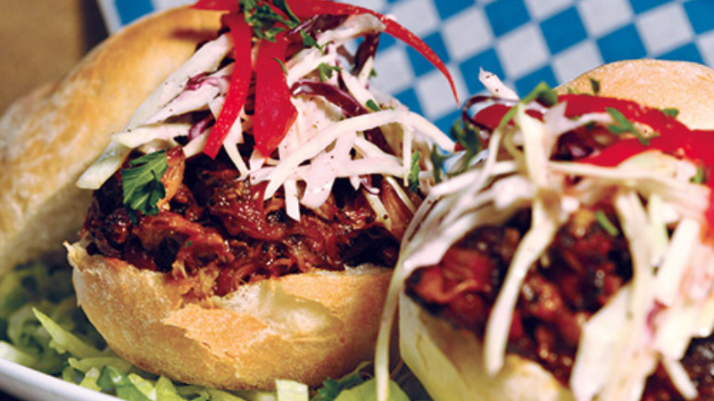 Two pulled meat sandwiches topped with red peppers, shredded cabbage, and fresh herbs, placed on a bed of lettuce.