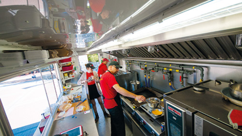 Inside view of a food truck with two staff members cooking and preparing food in the kitchen area, featuring kitchen equipment and appliances.