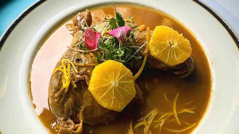 A plate with chicken served in sauce, garnished with orange slices, herbs, and edible flowers.
