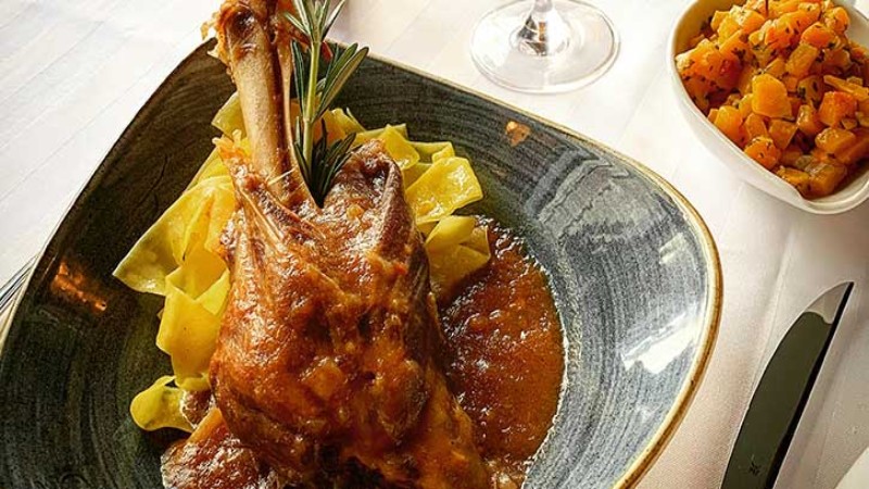 A plate with roasted lamb shank served with pasta, sauce, and a side of diced vegetables.
