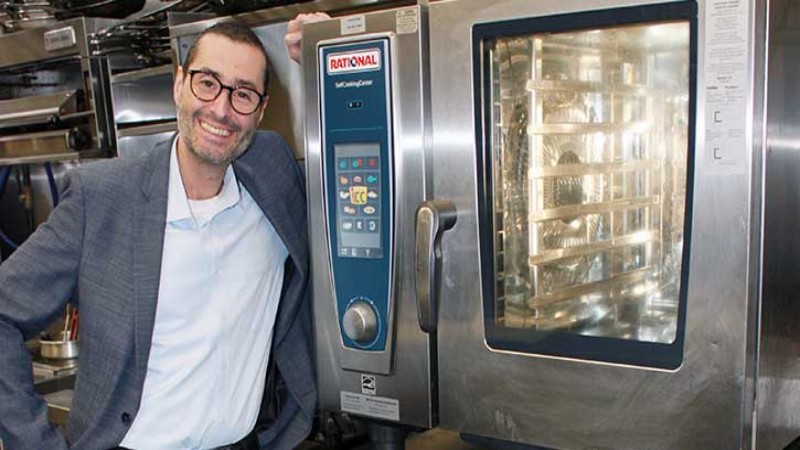 A man in a suit standing next to a Rational oven in a kitchen.