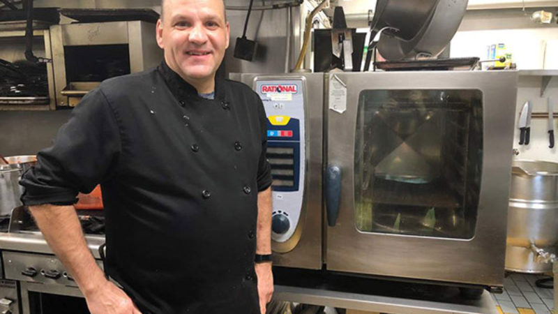 A chef in a black uniform standing beside a Rational oven in a kitchen.