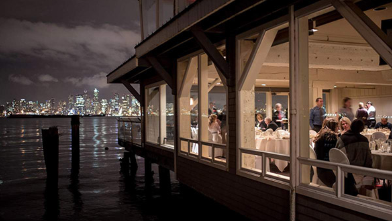 A view of a waterfront restaurant with diners inside, illuminated by city lights at night.