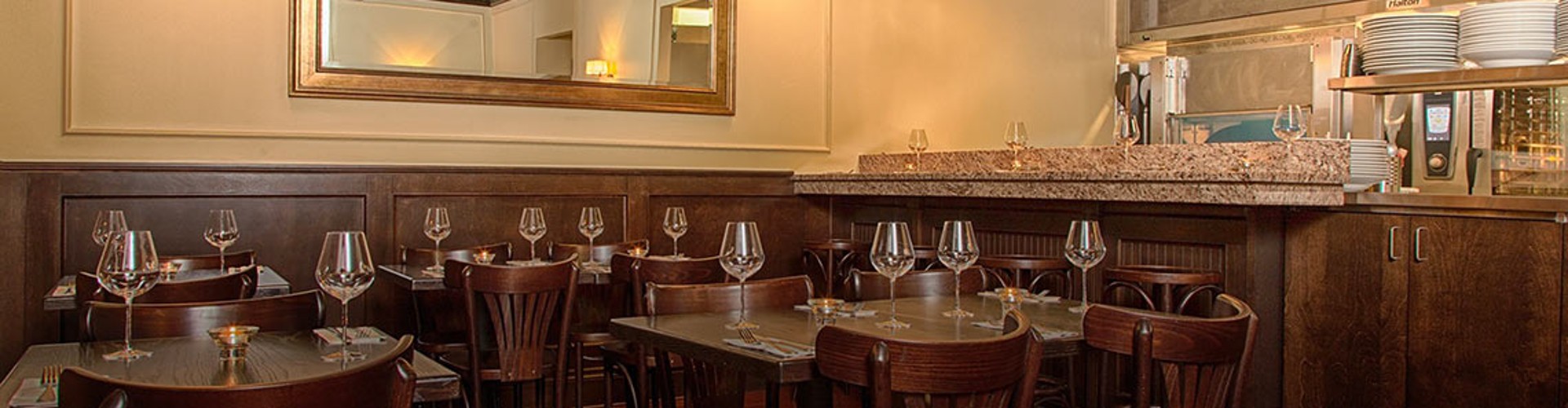 Elegant dining area with neatly arranged tables and chairs, each table set with wine glasses and plates. A well-lit bar with stools and a mirror spans the background.