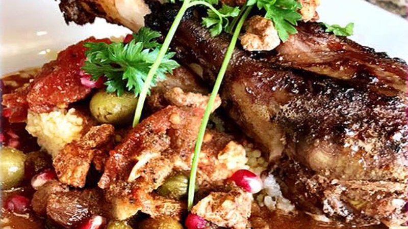 A plate featuring braised lamb shank served over couscous with tomatoes, olives, and garnished with parsley.