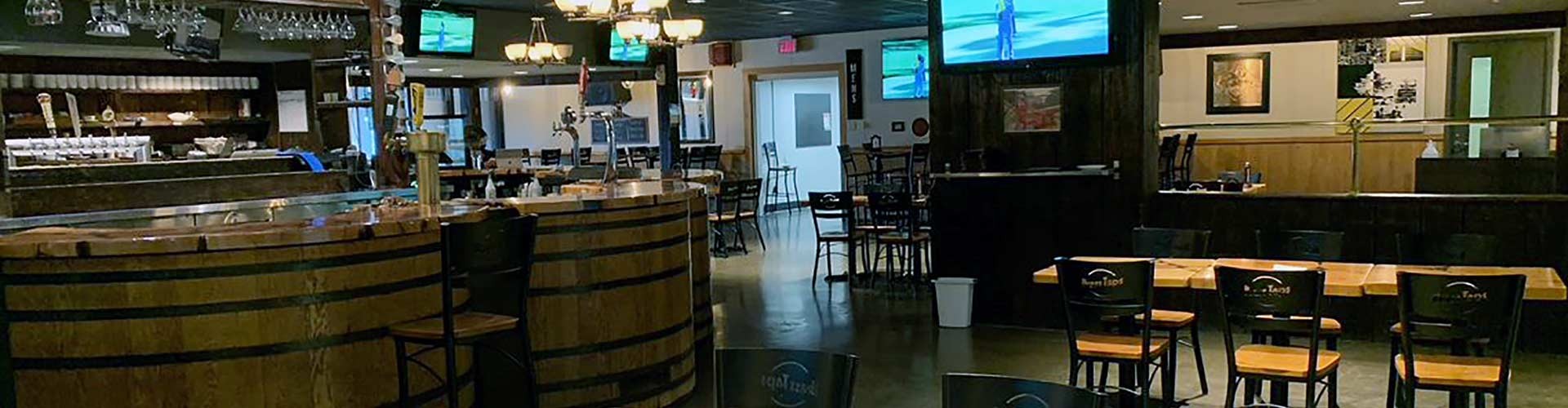 Interior of a bar featuring a well-stocked counter, bar stools, barrel tables, and multiple televisions displaying sports channels.