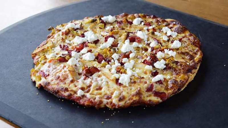 A pizza with toppings of corn, red peppers, and dollops of white cheese, served on a black slate board.