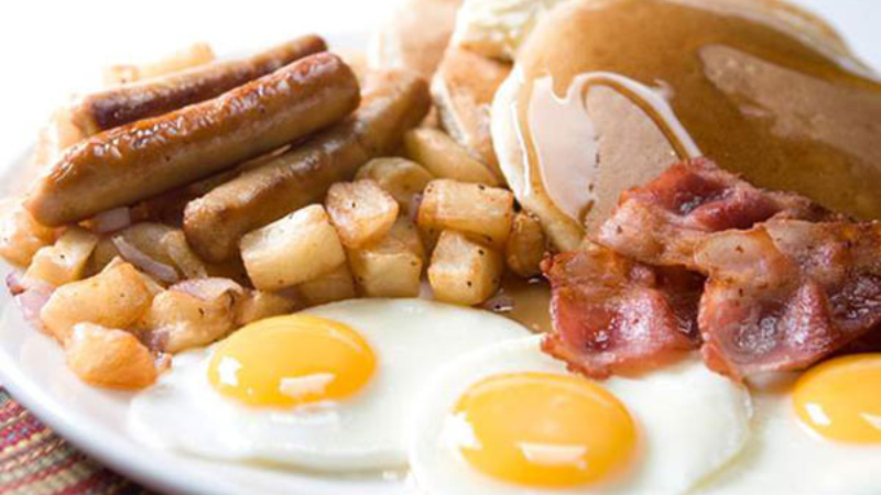 A plate of breakfast food featuring pancakes with syrup, bacon, sausages, diced potatoes, and two sunny-side up eggs.