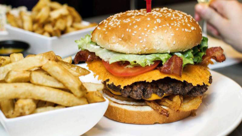 A close-up image of a cheeseburger with bacon, lettuce, and tomato, served on a sesame seed bun accompanied by a side of French fries.