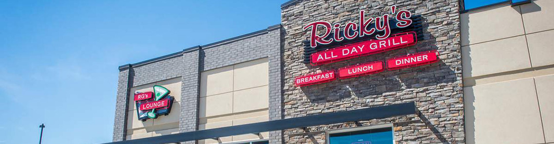 Exterior view of Ricky‘s All Day Grill restaurant featuring modern architecture with stone facades, large windows, and signs depicting the restaurant‘s name and logo. A parked car is visible in the foreground.