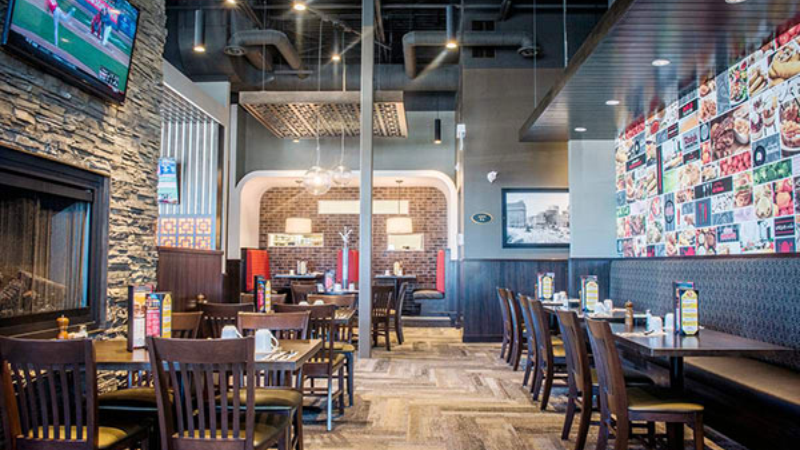 Interior of a sports-themed restaurant featuring multiple dining tables and chairs, a stone fireplace on the left, and a wall decorated with sports photographs. TVs displaying sports are mounted on the walls, and condiments are placed on each table.