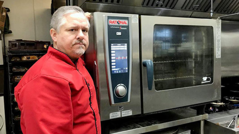 A person in a red chef jacket standing next to a Rational combi oven in a commercial kitchen.