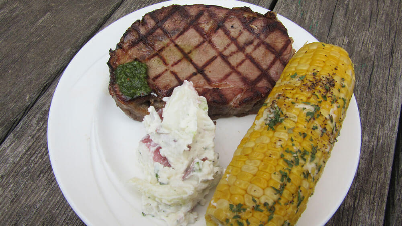 A plate of grilled steak with a grilled corn on the cob and a serving of creamy potato salad.
