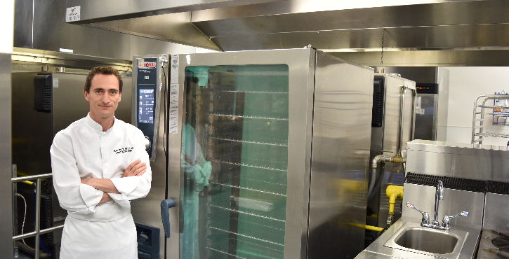 A chef standing in a commercial kitchen with arms crossed, next to a large RATIONAL cooking oven and kitchen equipment.