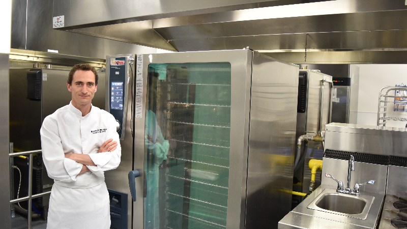 Un chef debout dans une cuisine professionnelle, bras croisés, à côté d‘un grand four RATIONAL et des équipements de cuisine.