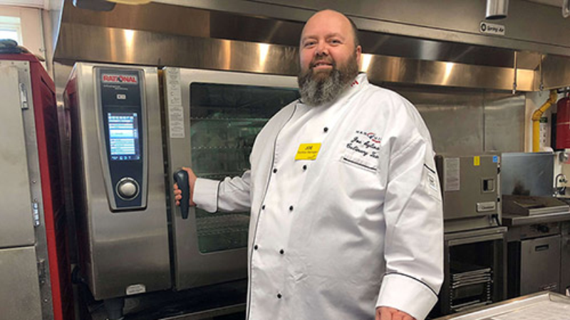 A chef standing in front of a Rational oven, wearing a white chef coat and a name tag in a professional kitchen.