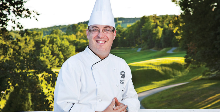 A chef in a white coat and hat, smiling and standing outdoors with a green landscape in the background.