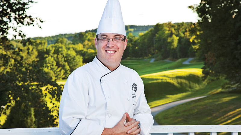 Un chef en manteau et toque blancs, souriant et debout à l‘extérieur avec un paysage verdoyant en arrière-plan.