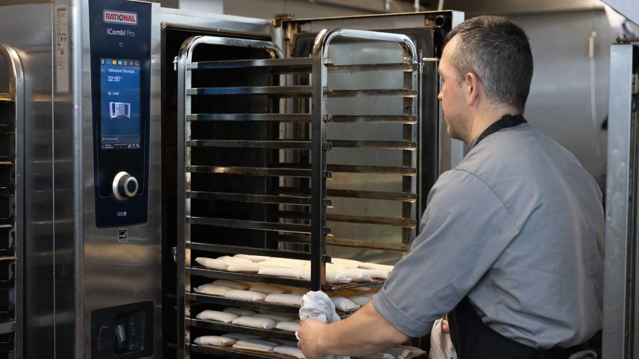 A baker loads a rack of dough pieces into a RATIONAL iCombi Pro combi oven in a production kitchen.