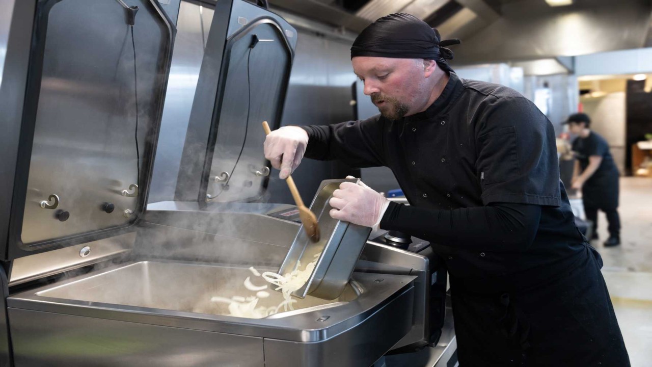 Chef stirs sliced onions in a steaming RATIONAL iVario Pro tilting pan in a busy professional kitchen.