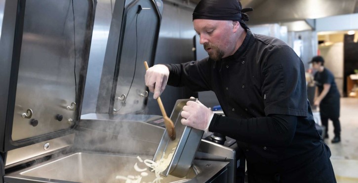 Chef stirs sliced onions in a steaming RATIONAL iVario Pro tilting pan in a busy professional kitchen.