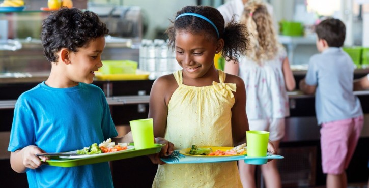 Two smiling children carry trays with vegetables and drinks in a school canteen.