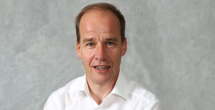 Portrait of a man in a white shirt speaking against a light grey background.