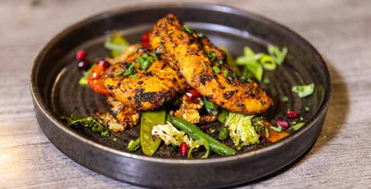 A plate of food from IndiYang. Tandoori chicken sits on top of a bed of grilled colourful vegetables, garnished with coriander, spring onion and pomegranate. The food is on a black frost plate