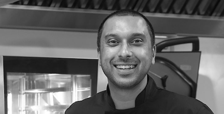 Rehan Uddin smiling in a kitchen environment, wearing a chef’s jacket.