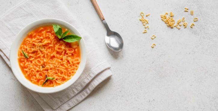 An overhead view of a white bowl of tomato alphabet soup, with fresh basil leaves, sits on a white tea towel, next to a spoon, on a marble work surface. There is some spare dried alphabet spaghetti letters sprinkled onto the work surface on the side.