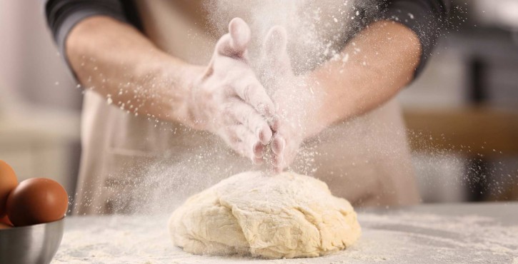 A baker slaps their hands together causing a cloud of flour to fall on top of some bread dough.