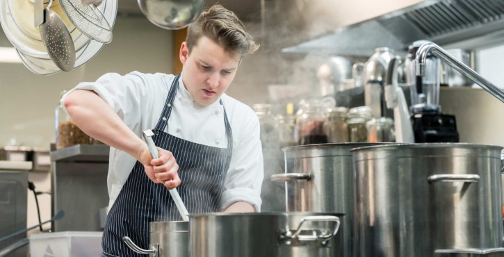 A young, white male chef in an apron stirs a pot of boiling liquid, in a restaurant kitchen. There is 3 big pots in front of him and lots of kitchen utensils hanging above him.