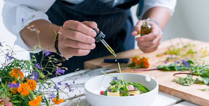 A chef uses a pipette to add some oil onto a beautifully plated fine dining dish with pasta and small purple flowers.