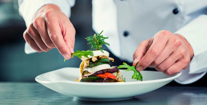A chef puts the finishing touches on his dish. He adds rocket leaves to a grilled vegetable tower, on a white plate.