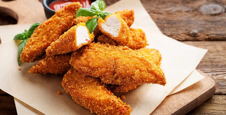 Some crispy baked golden chicken nuggets served in a pile on a wooden board with a small pot of ketchup.