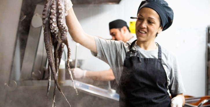 A female chef in a black hat and black apron, lifts a raw octopus from a gastro of other octopus and holds it over the iVario Pro, ready to cook.