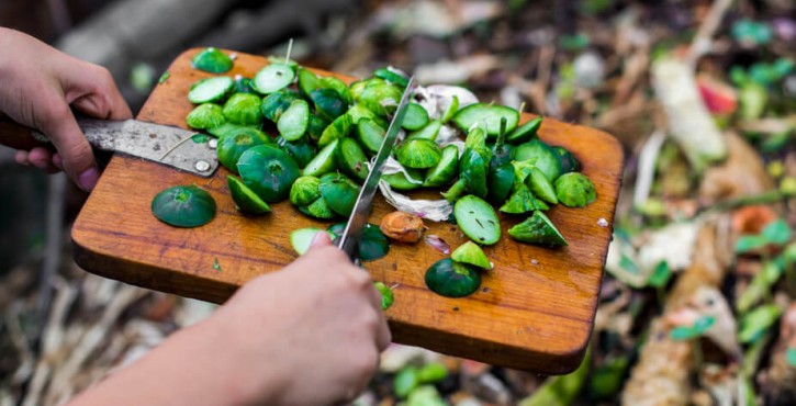 An overhead photo of a person holding a chopping board over a compost pile, scraping the ends of courgettes and other vegetable waste onto the top of the pile with a knife.