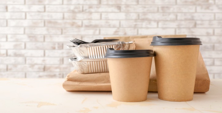 2 cardboard coffee cups, with black lids sit on a work bench in front of a brown paper bag on which sits 2 aluminium containers with 2 forks on top, all in front of a grey brick wall.