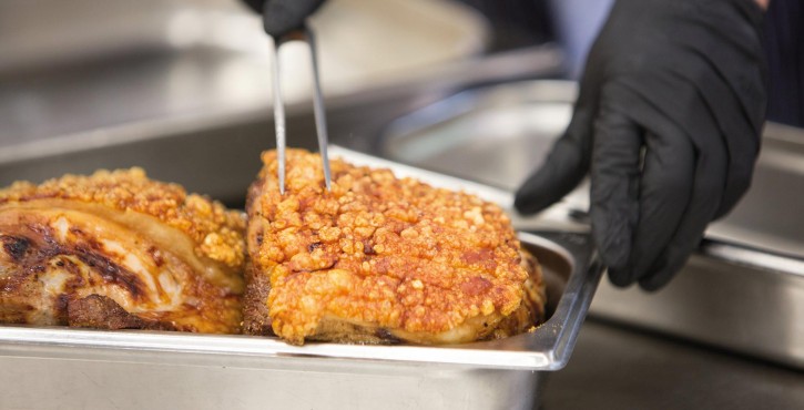 A pair of black gloved hands put a carving fork into a roasted pork join with golden crackling on top, which sits in a tray.