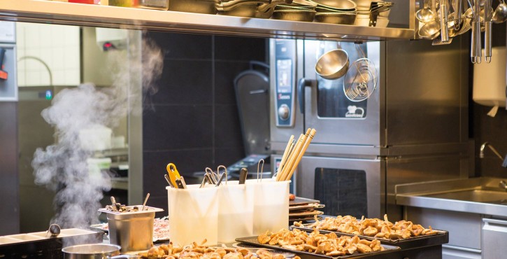 A restaurant kitchen with no staff, with a RATIONAL oven in the background. Shelves are full of ladles, spoons and other equipment. Food is ready to be served, sitting on the counter.
