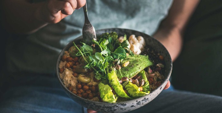 A dark image of someone holding a grey bowl full of hummus topped with avocado, beans, lettuce and greens. They are putting their fork into it to start eating.